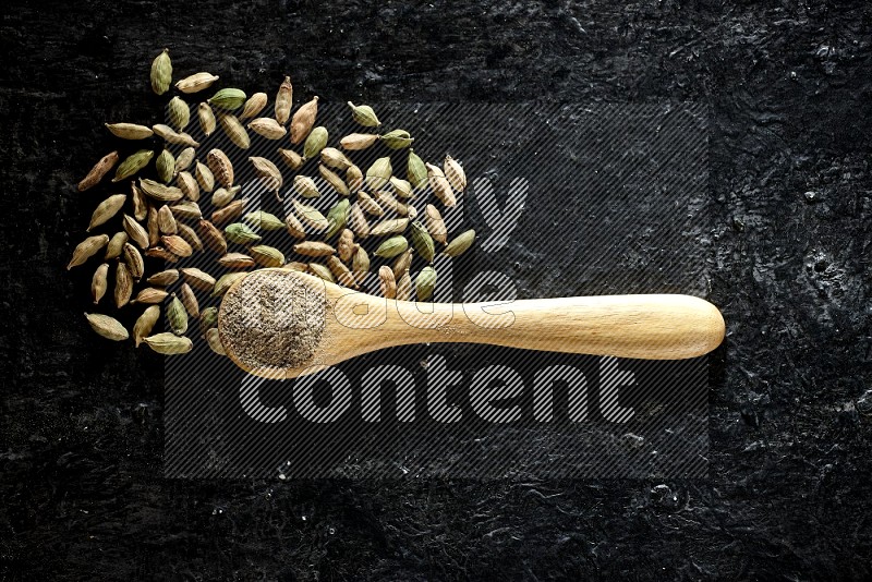 A wooden spoon full of cardamom powder and cardamom seeds next of it on textured black flooring