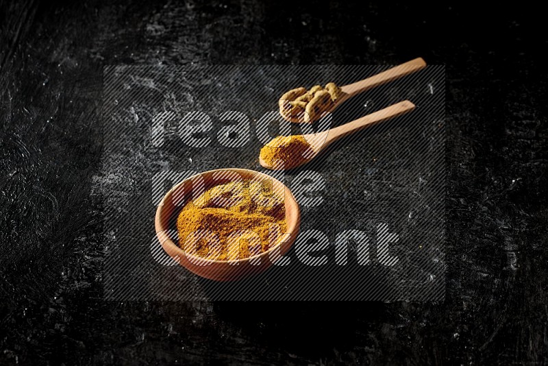 A wooden bowl full of turmeric powder and 2 wooden spoons full of dried turmeric finger and turmeric powder on textured black flooring