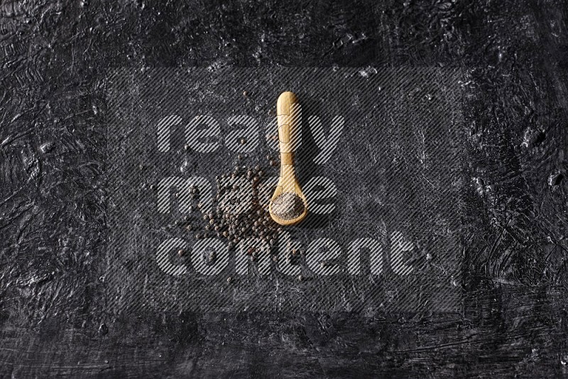 A wooden spoon full of black pepper powder and black pepper beads spread on a textured black flooring