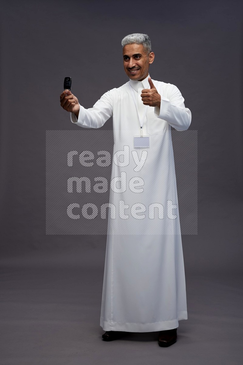 Saudi man wearing thob with neck strap employee badge standing holding key car on gray background