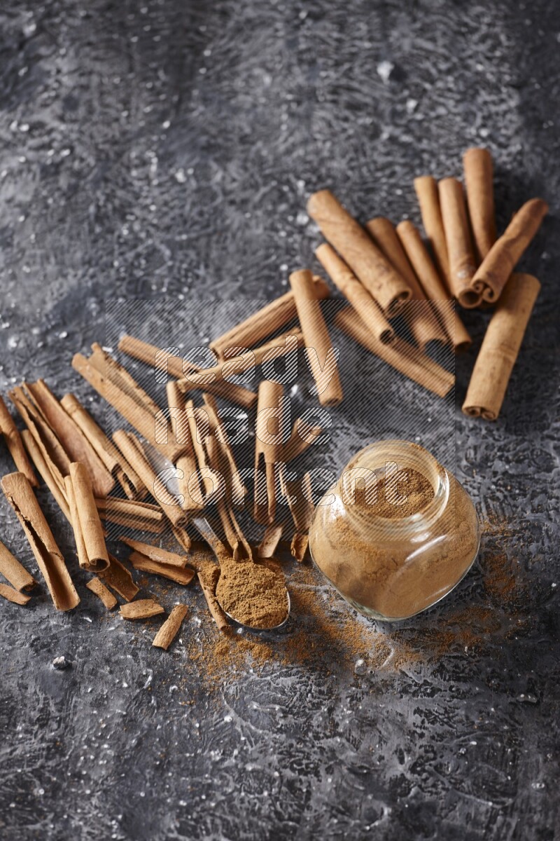 Herbal glass jar and a metal spoon full of cinnamon powder surrounded by cinnamon sticks on textured black background