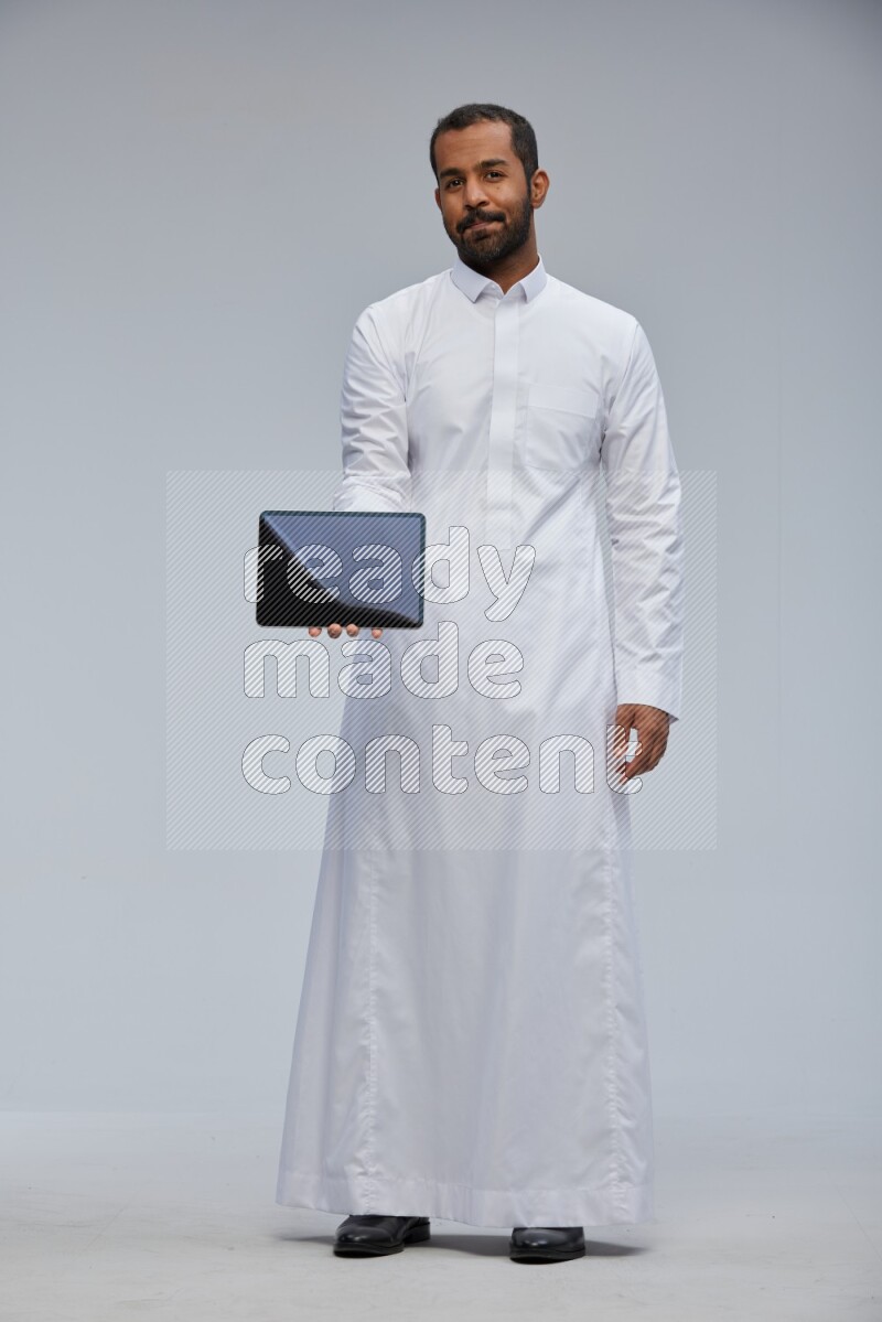 Saudi man wearing Thob standing showing tablet to camera on Gray background