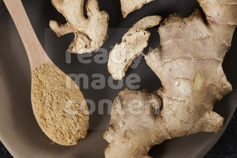 A wooden spoon full of ground ginger powder with fresh and dried ginger, all on a pottery plate on black background