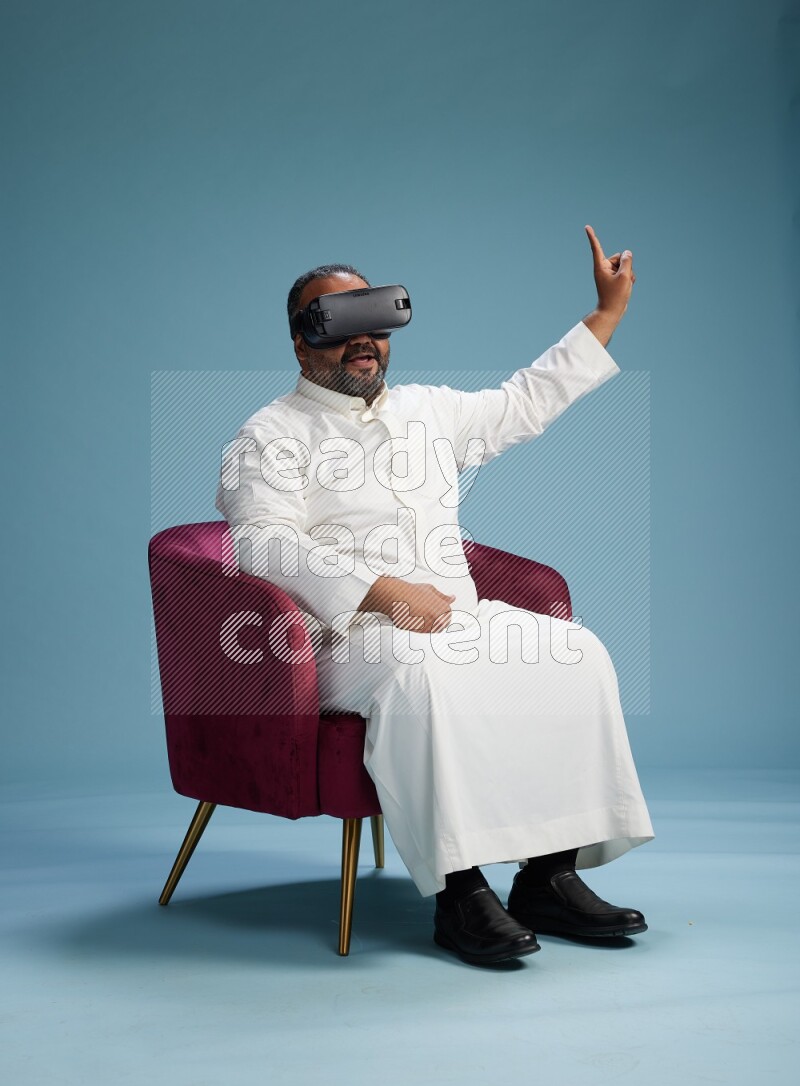 Saudi Man without shimag sitting on chair  gaming with VR on blue background