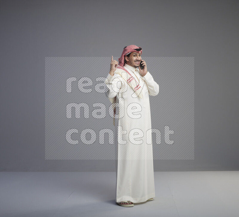 A Saudi man standing wearing thob and red shomag talking on phone on gray background