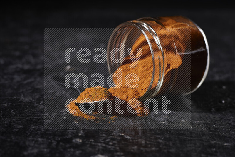 A glass jar full of ground paprika powder flipped with some spilling powder on black background