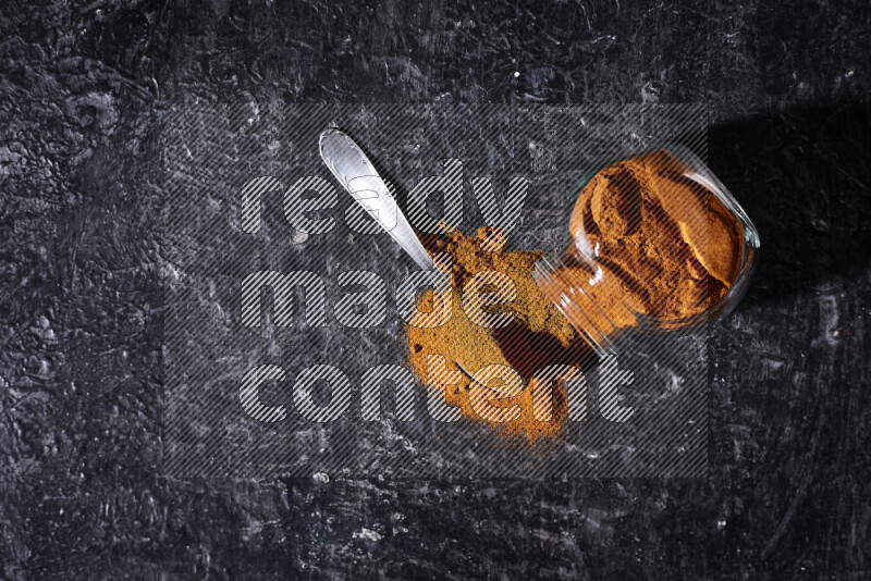 A glass jar full of ground paprika powder flipped with some spilling powder on black background