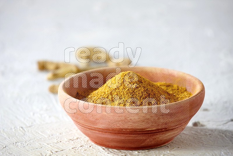 A wooden bowl full of turmeric powder with dried whole fingers on textured white flooring