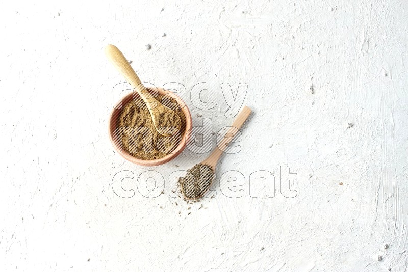 A wooden bowl and 2 wooden spoons full of cumin powder and cumin seeds on textured white flooring