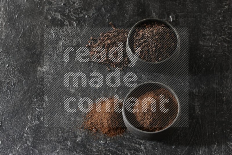 2 Black pottery bowls full of cloves and the other full of cloves powder on textured black flooring