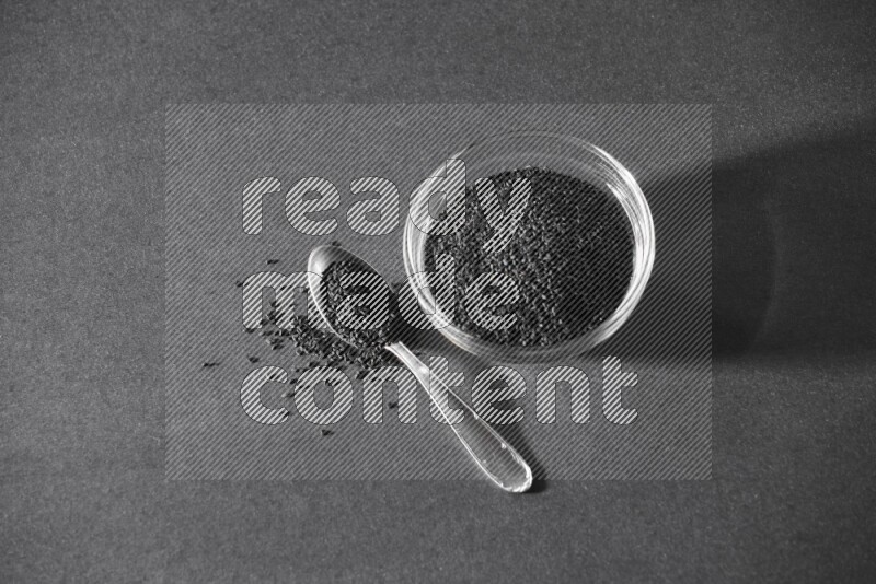 A glass bowl full of black seeds with a metal spoon full of the seeds on a black flooring