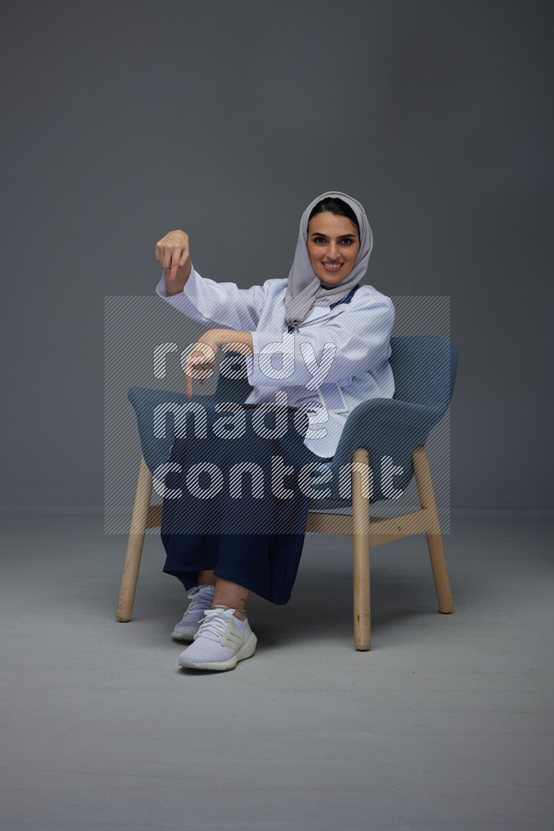 A doctor wearing a light gray head scarf setting on a dark grey chair and using a phone eye level on a grey background