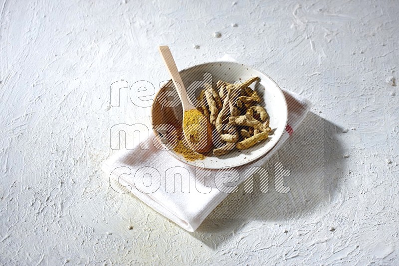 A plate filled with dried turmeric whole fingers and a wooden spoon full of turmeric powder on a textured white flooring