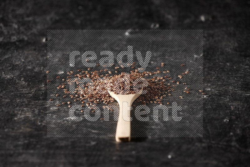 A wooden spoon full of flaxseeds and surrounded by seeds on a textured black flooring