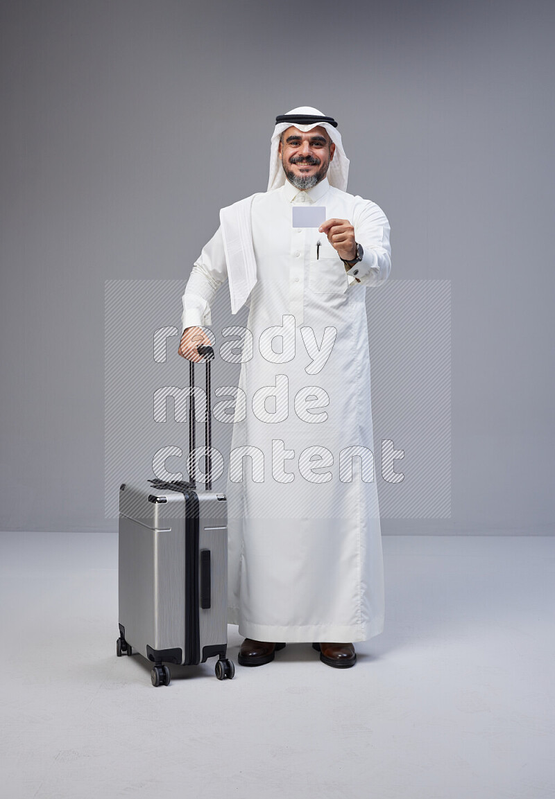 Saudi man wearing Thob and white Shomag standing holding Travel bag and ATM card on Gray background