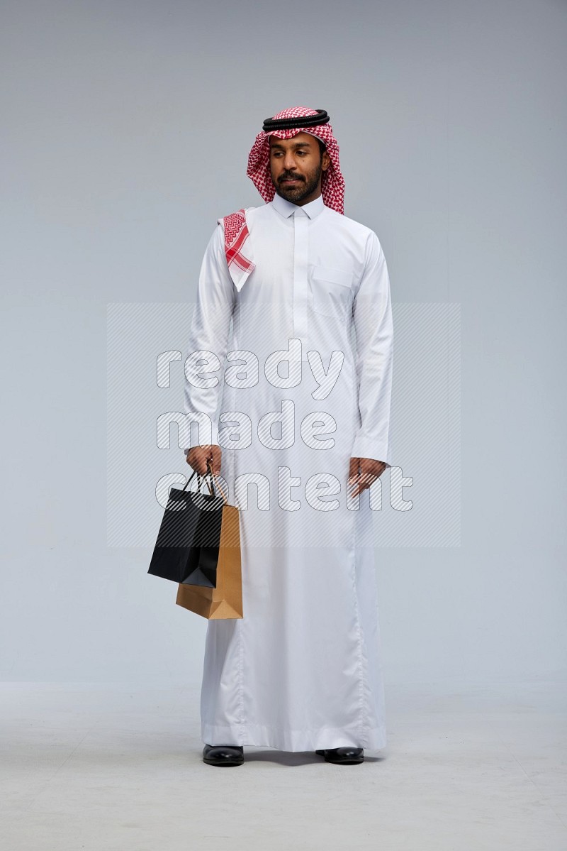 Saudi man Wearing Thob and shomag standing holding shopping bag on Gray background
