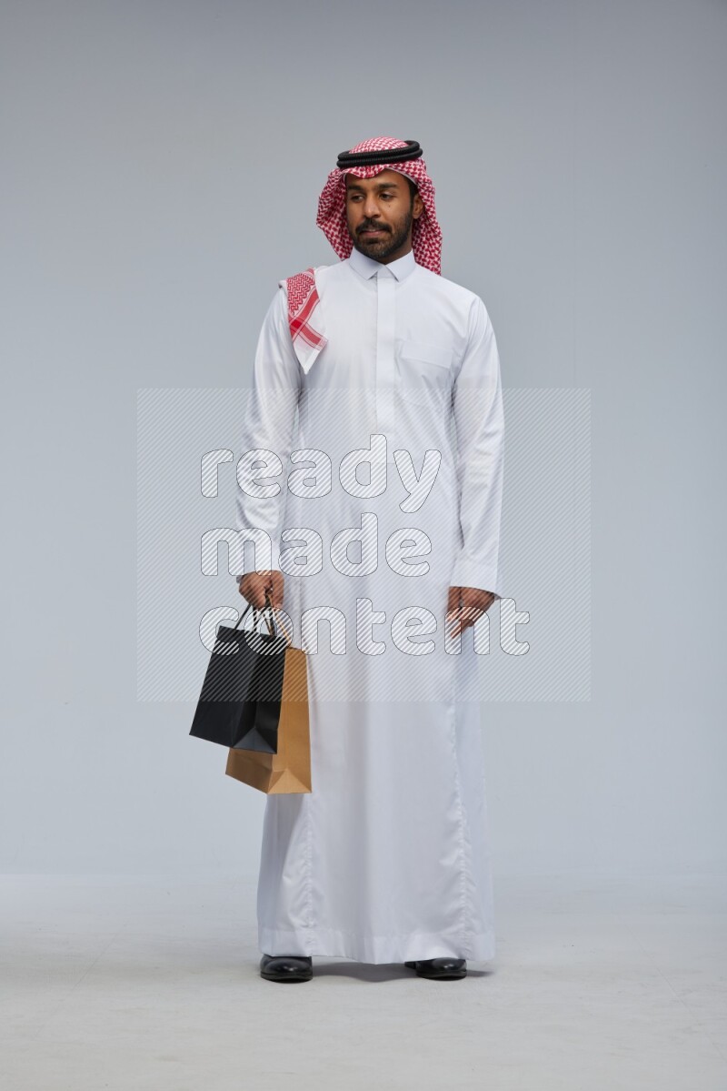 Saudi man Wearing Thob and shomag standing holding shopping bag on Gray background