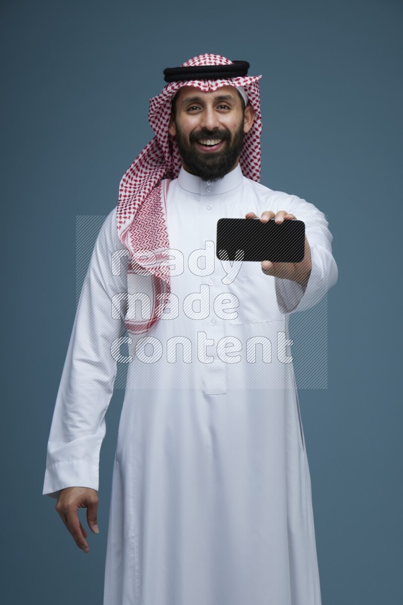 A man showing his phone Screen in a blue background wearing Saudi Thob with Shomag