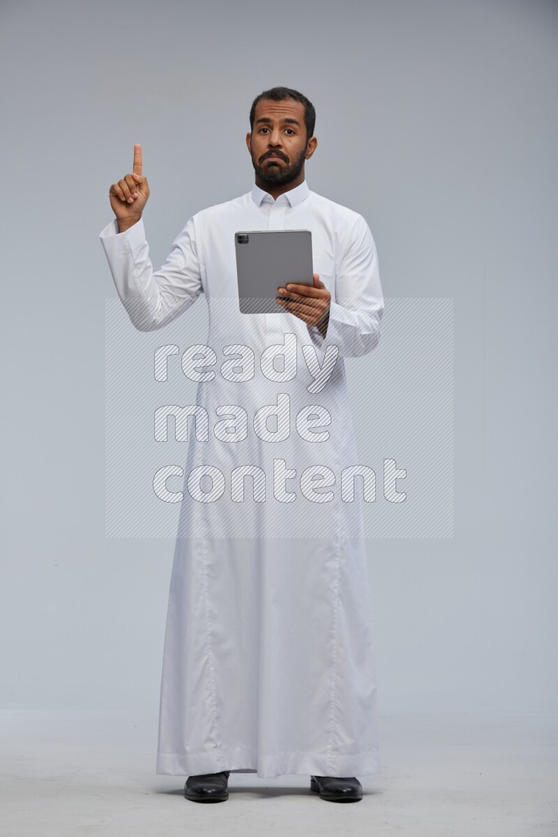 Saudi man wearing Thob standing working on tablet on Gray background