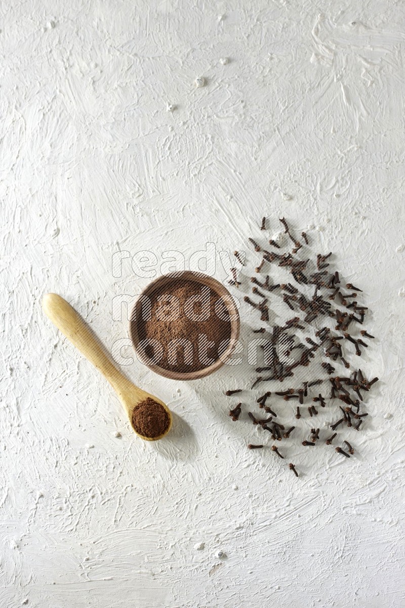 A wooden bowl and wooden spoon full of cloves powder with cloves spread on textured white flooring