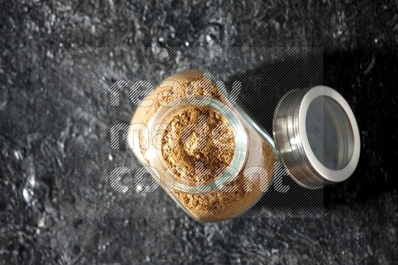 A glass spice jar full of allspice powder on a textured black flooring