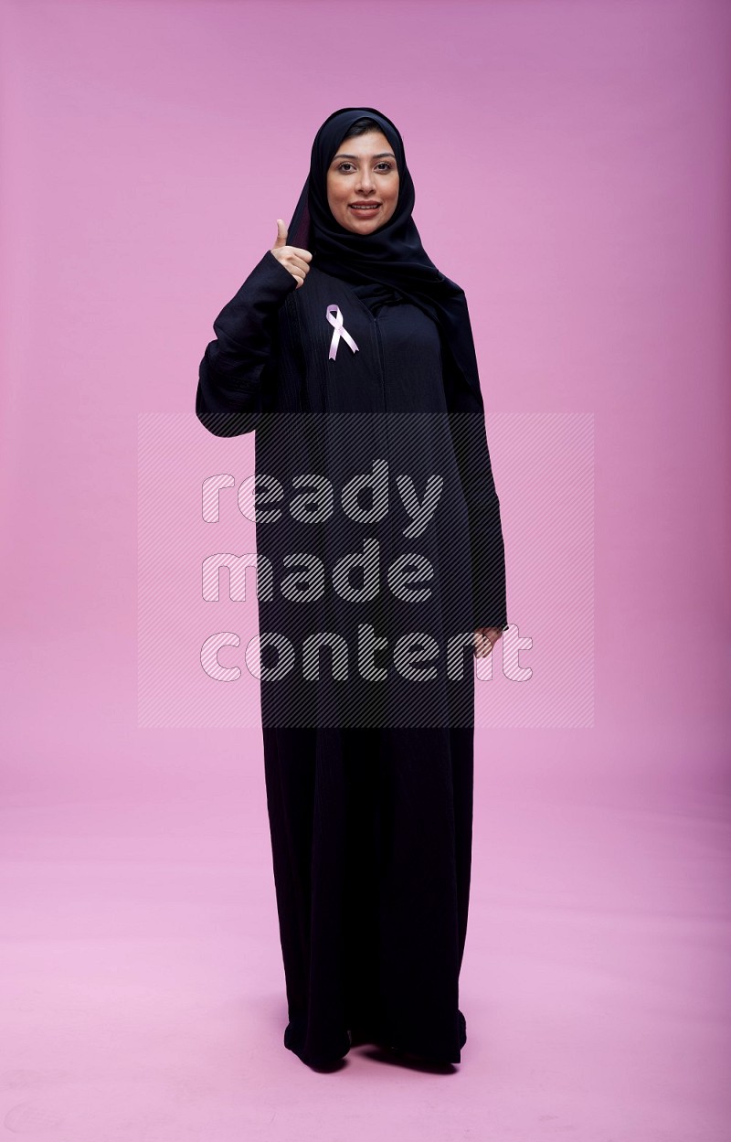 Saudi woman wearing pink ribbon on Abaya standing interacting with the camera on pink background