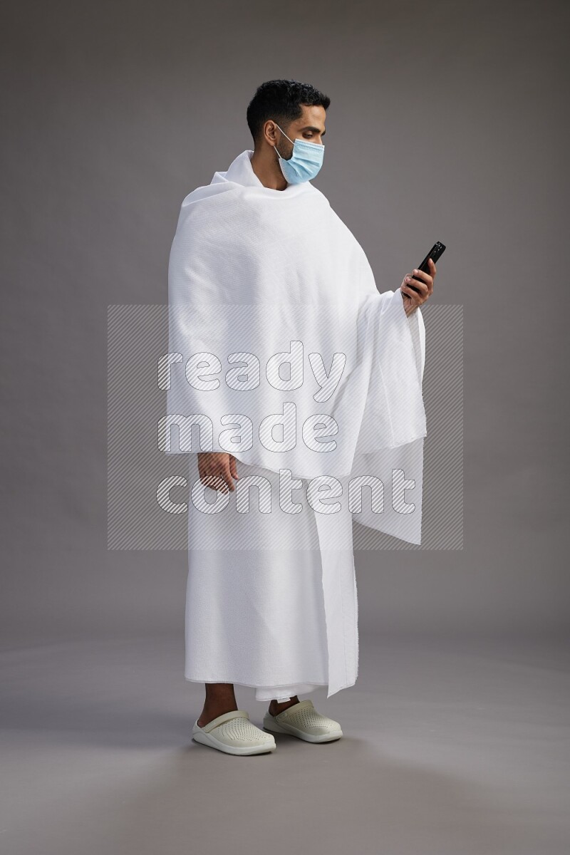 A man wearing Ehram with face mask Standing texting on phone on gray background