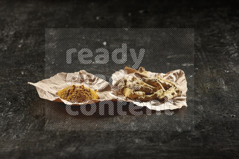 Turmeric powder and dried turmeric whole fingers in 2 crumpled pieces of paper on textured black flooring