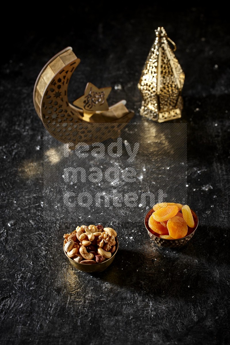 Nuts in a metal bowl with dried apricots beside golden lanterns in a dark setup