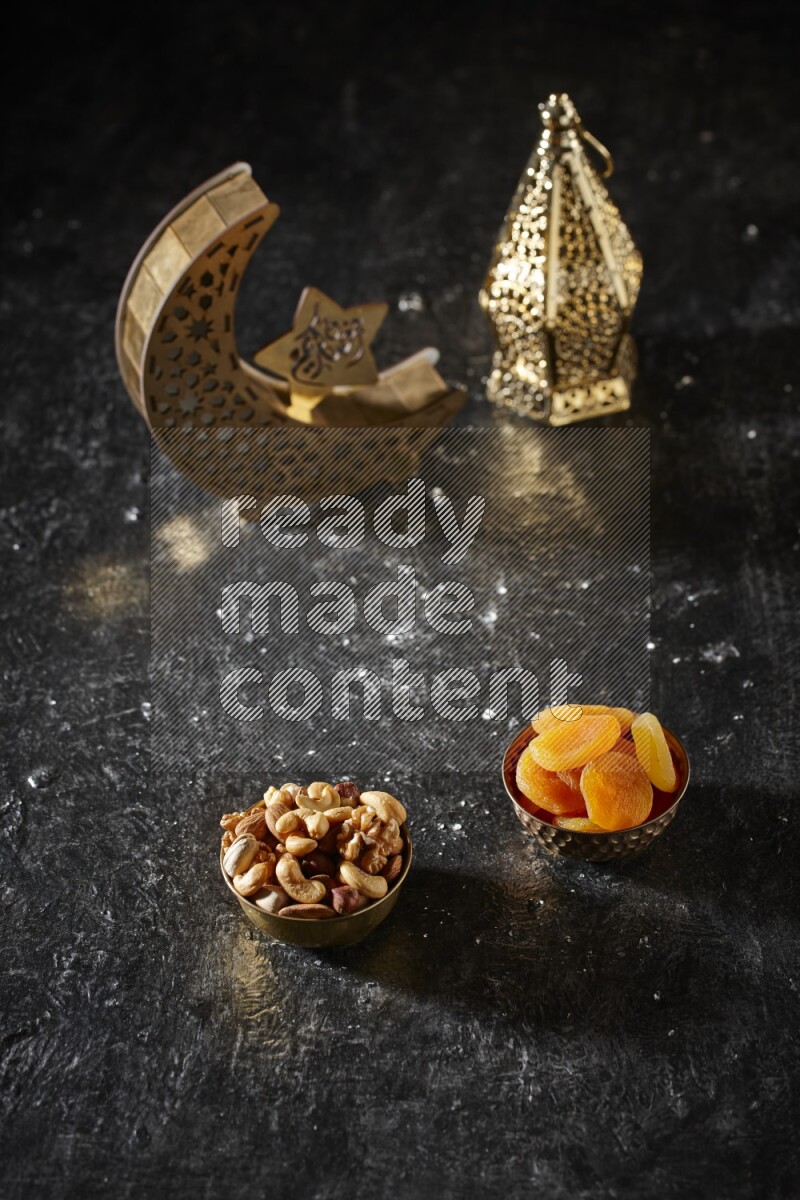 Nuts in a metal bowl with dried apricots beside golden lanterns in a dark setup
