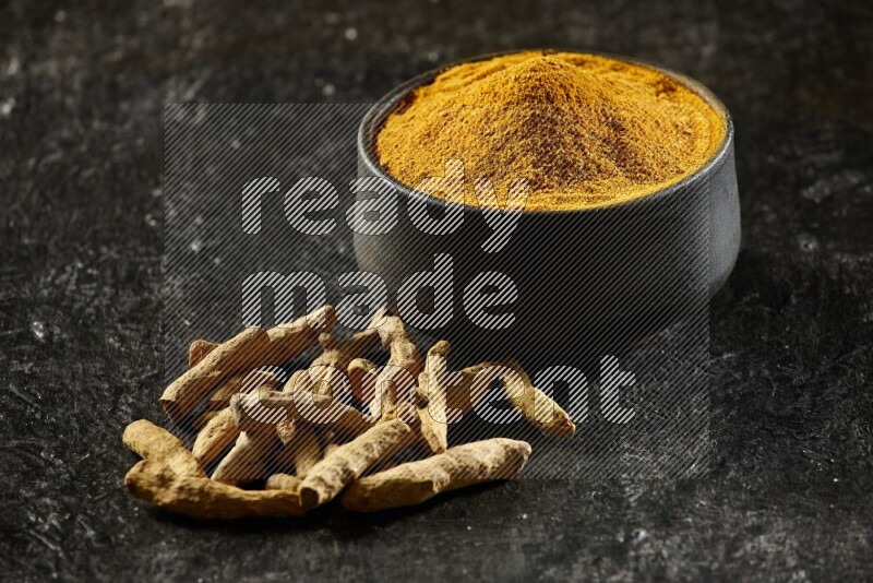 A black pottery bowl full of turmeric powder and dried turmeric whole fingers next of it on textured black flooring