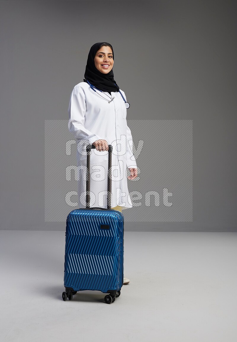 Saudi woman wearing lab coat with stethoscope standing holding Travel bag on Gray background