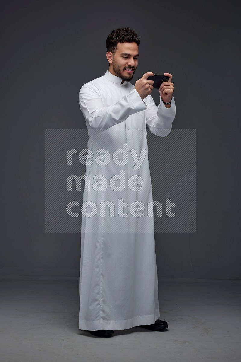 A Saudi man wearing Thobe and playing using his phone horizontally playing video games while standing and making different poses eye level on a gray background