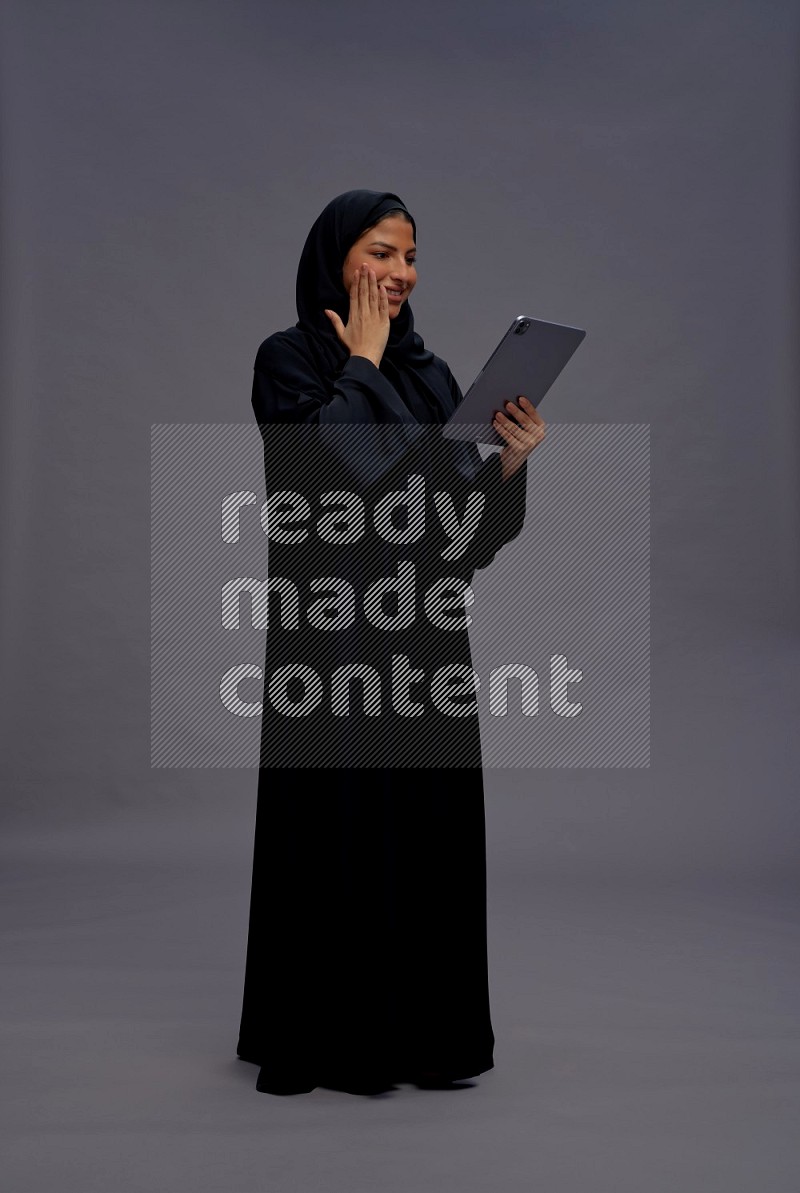 Saudi woman wearing Abaya standing working on tablet on gray background