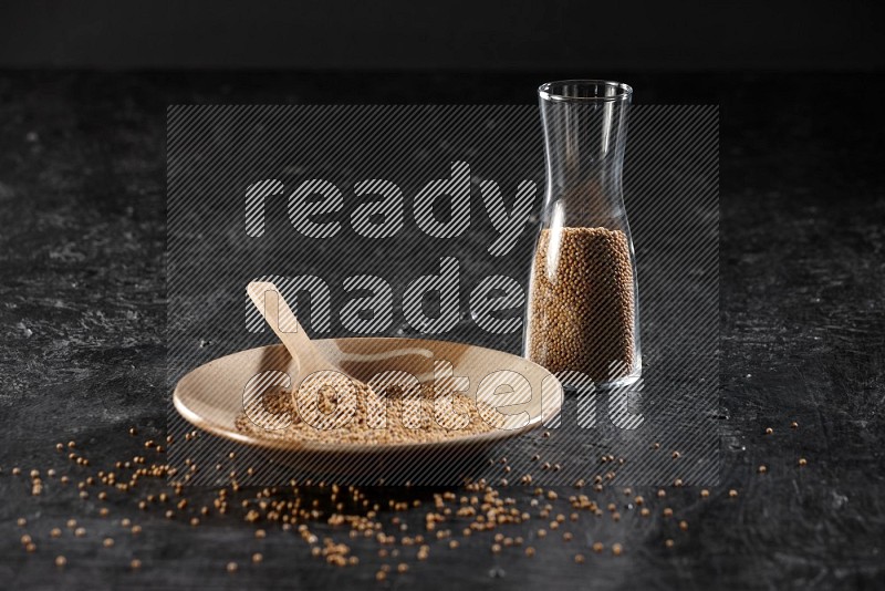 A beige pottery plate full of mustrad seeds and a wooden spoon in it with a glass jar filled with the seeds on a textured black flooring