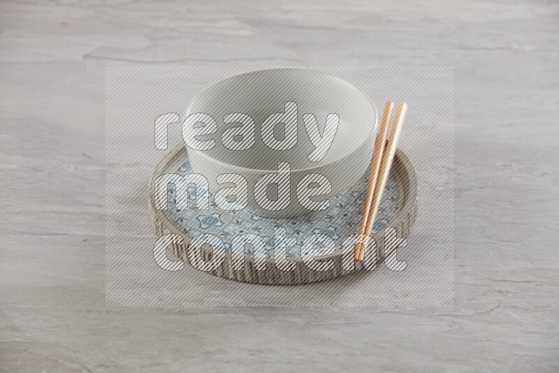 white pottery round bowl on top of multi color round ceramic plate and chopsticks, on grey textured countertop