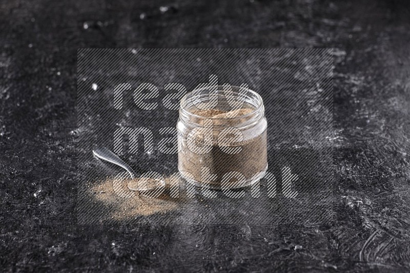 بودرة الفلفل الأسود في وعاء زجاجي مع ملعقة معدنية مليئة بالبودرة علي خلفية سوداء