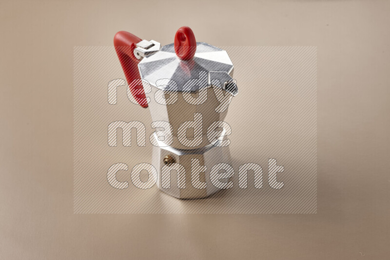 A moka pot with red handle surrounded by roasted coffee beans on beige background