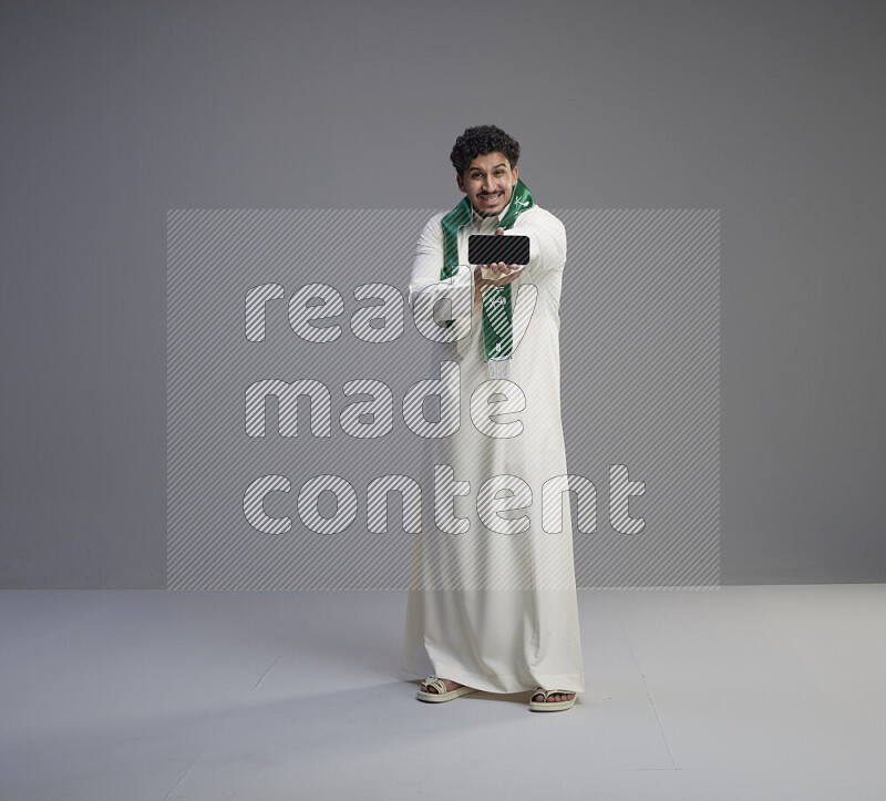 A Saudi man standing wearing thob and Saudi flag scarf showing phone to camera on gray background