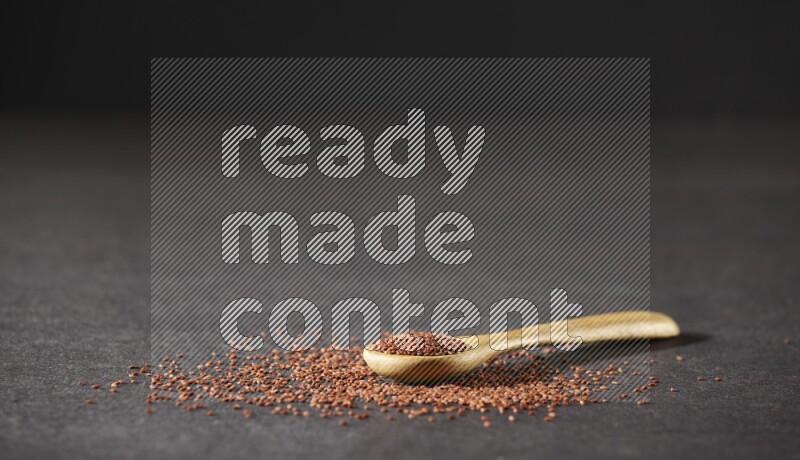 A wooden spoon full of garden cress seeds and seeds spread beside it on a black flooring