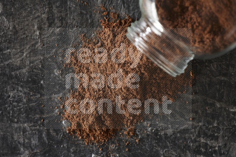 A flipped glass spice jar full of cloves powder and powder came out of it on textured black flooring