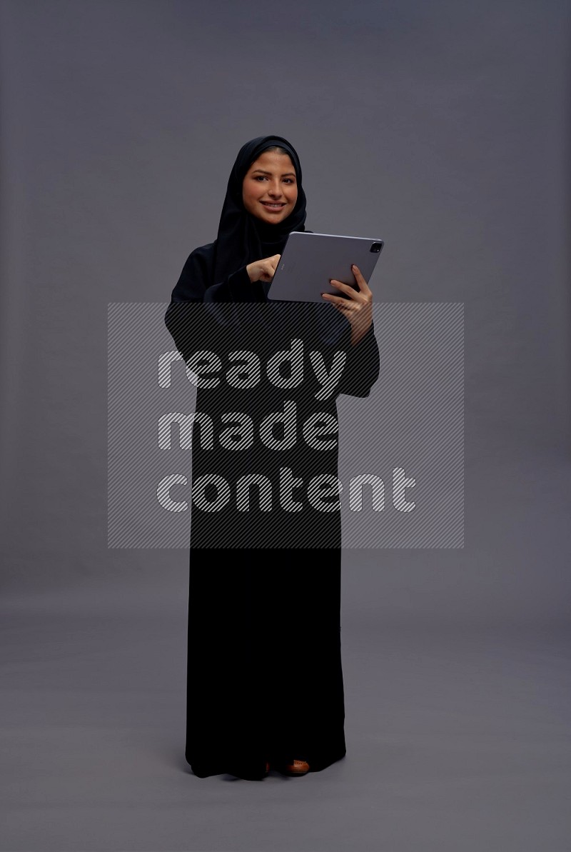 Saudi woman wearing Abaya standing working on tablet on gray background