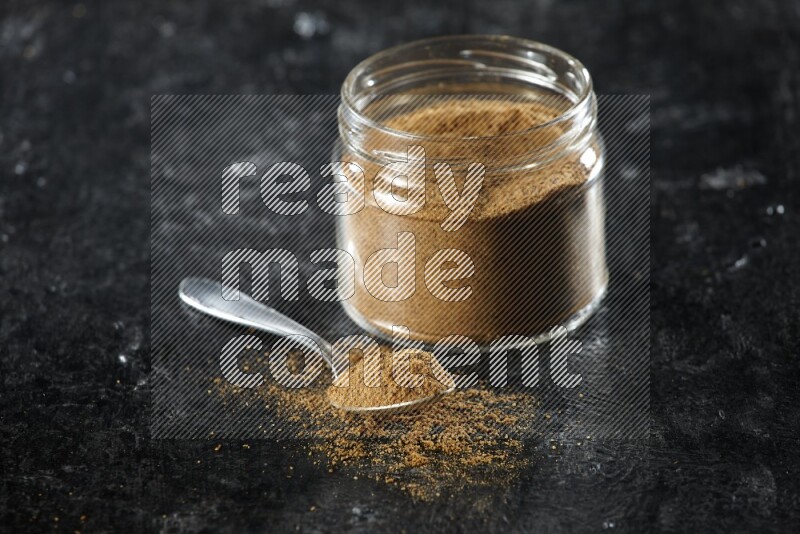 A glass jar and a metal spoon full of cumin powder on a textured black flooring