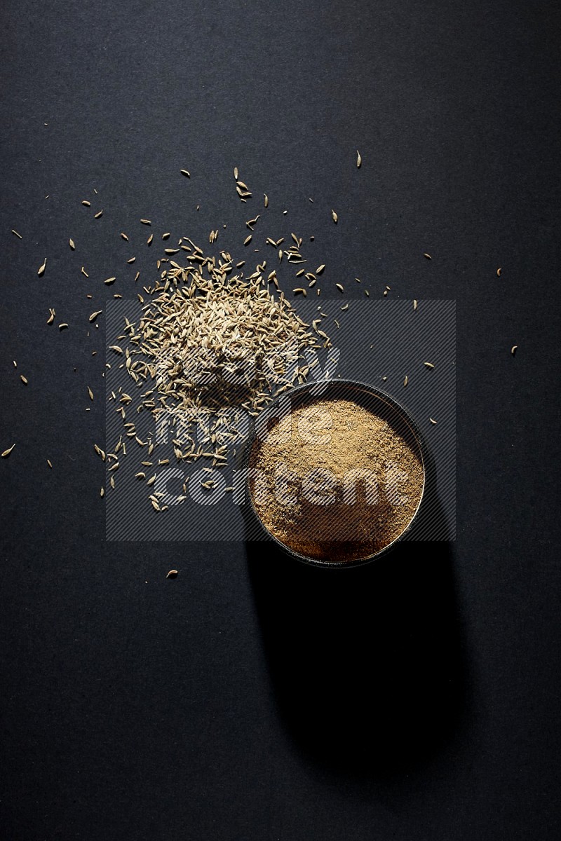 A black pottery bowl full of cumin powder and spreaded cumin seeds on a black flooring