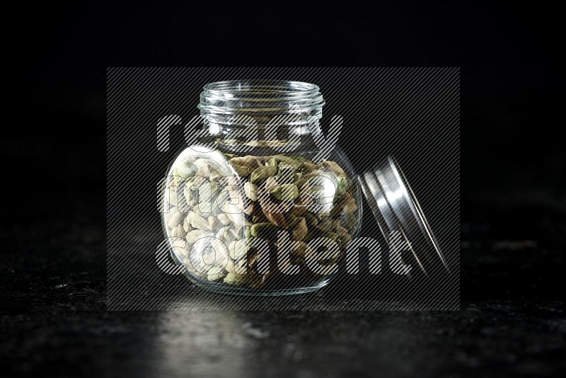A glass spice jar full of cardamom seeds on textured black flooring