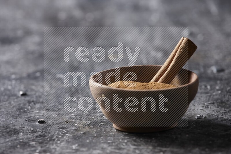 Wooden bowl full of cinnamon powder and a cinnamon stick on a textured black background