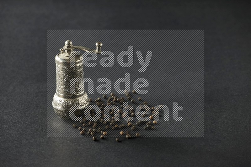A turkish metal pepper grinder and spreaded black pepper beads on a black flooring