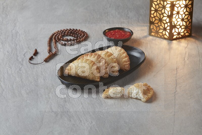 Four fried sambosas in an oval shaped black plate, beside a cut cheese sambosa, a brown misbaha and a golden lantern on a gray background