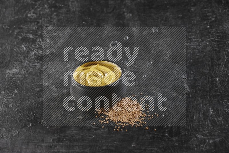 A black pottery bowl full of mustard paste with mustard seeds underneath on textured black flooring