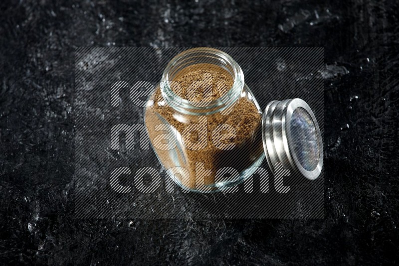 A glass spice jar full of cumin powder on a textured black flooring